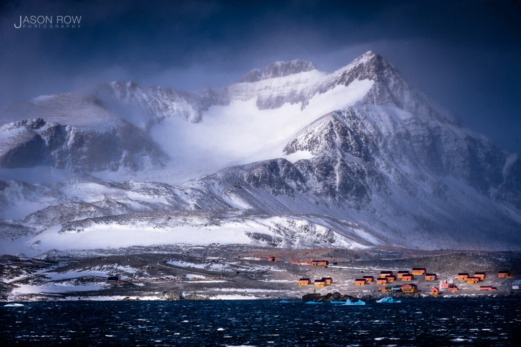 A stormy mountainous scene at the tip of the Antarctic peninsular. An Antarctic research base is seen at the foot of the mountain