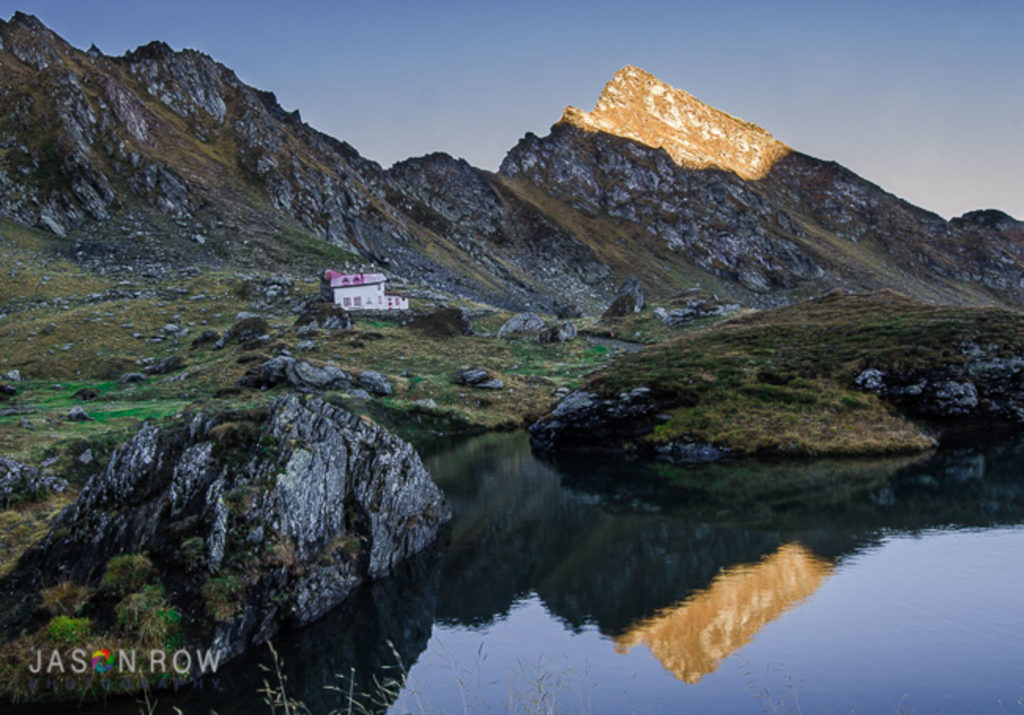 Transfagarasan Pass in Romania. A lake is overshadowed by mountain peaks. The sun just casting its light on the top of the peaks