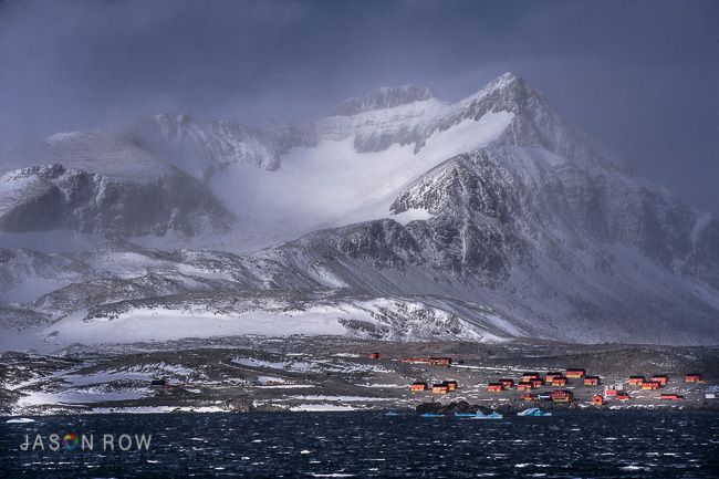 A stormy mountainous scene at the tip of the Antarctic peninsular. An Antarctic research base is seen at the foot of the mountain