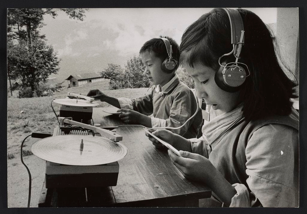 Photograph shows children seated at table outdoors, using 