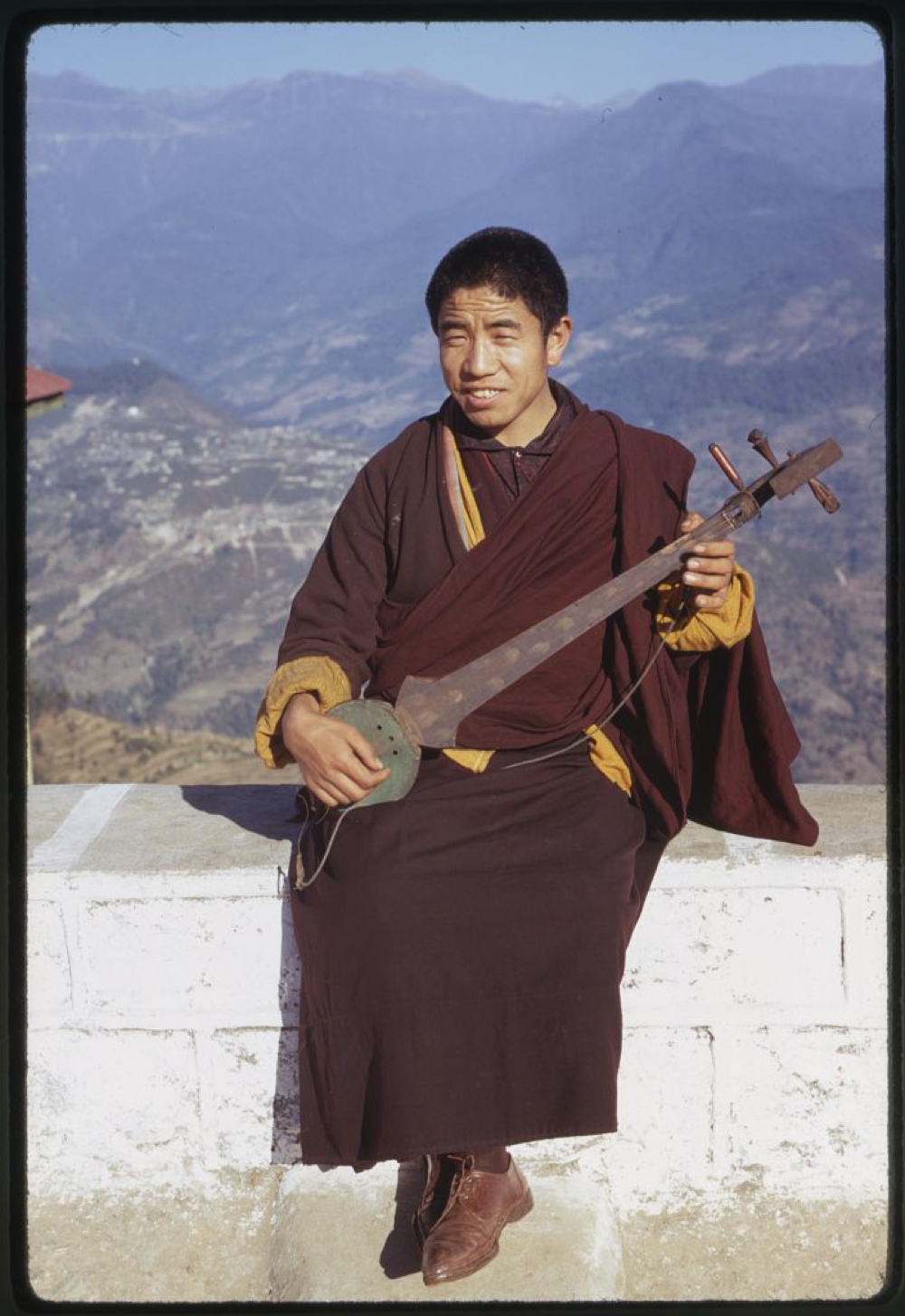 Photograph shows a monk, seated on a wall, playing a lute, Rumtek, Sikkim.