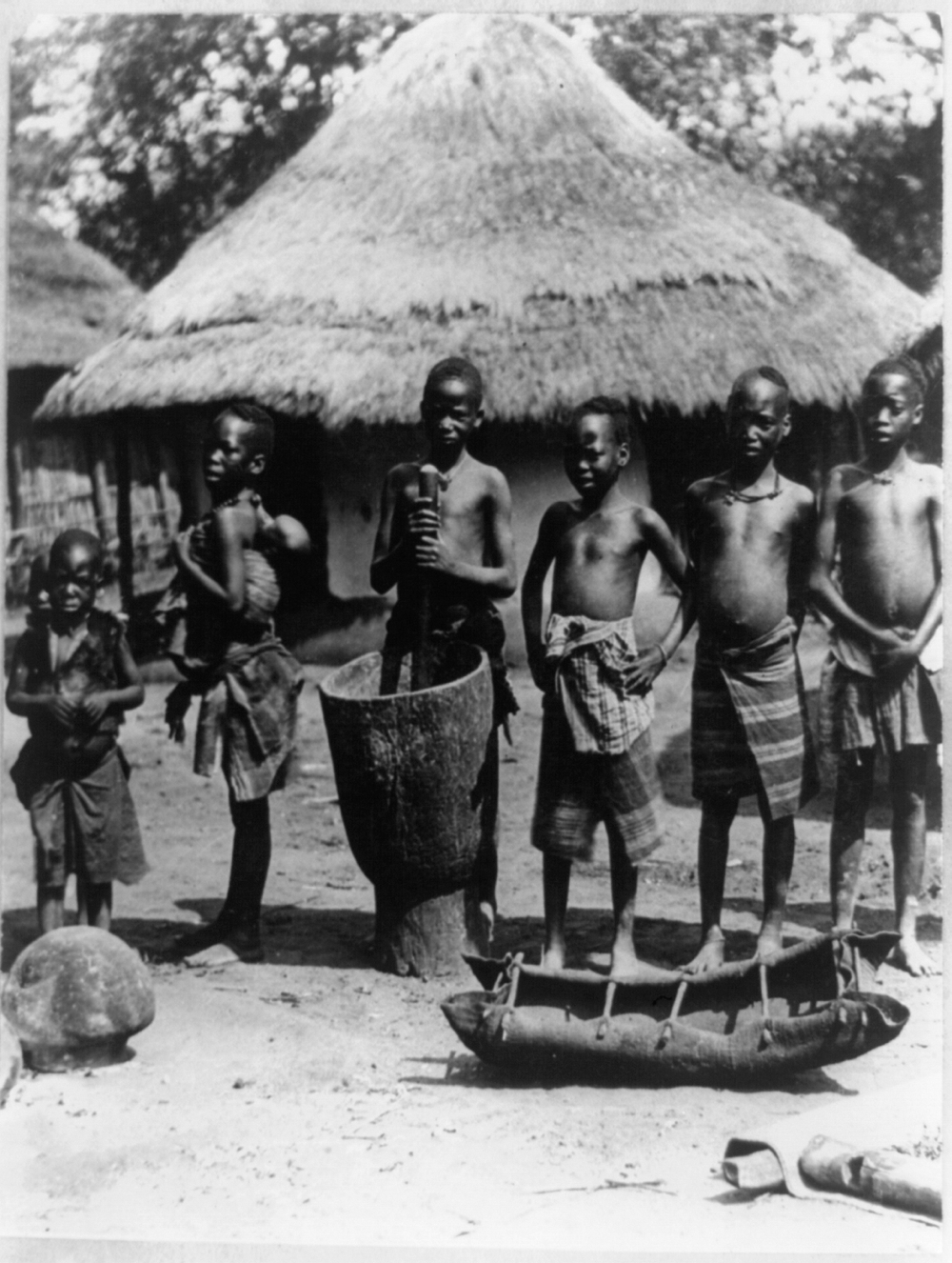 Seven native children posed in front of thatched-roof house, Portuguese East Africa