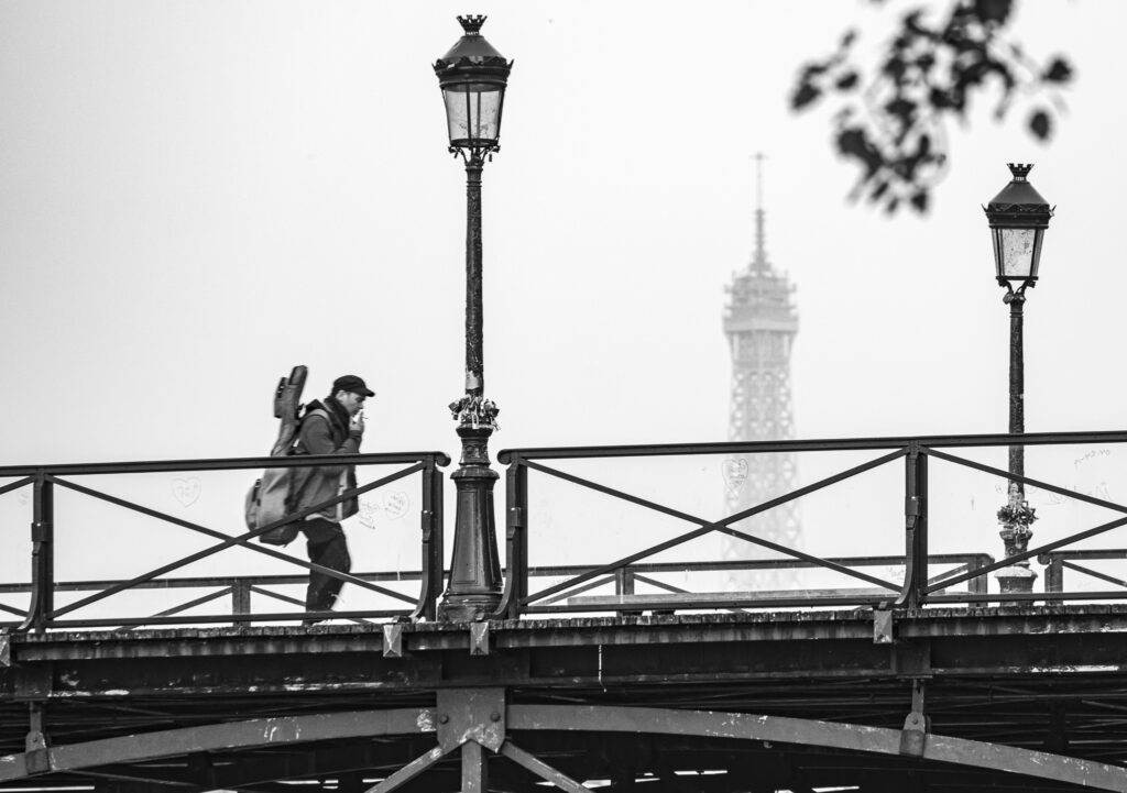 Musician walks across a Paris bridge with the Eifell Tower behind 