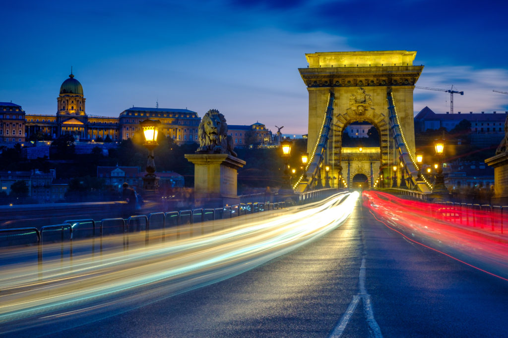 Twilight shot of traffic and light trails crossing the Chain Bridge in Budapest. The city lights are on and Buda Castle is in the background