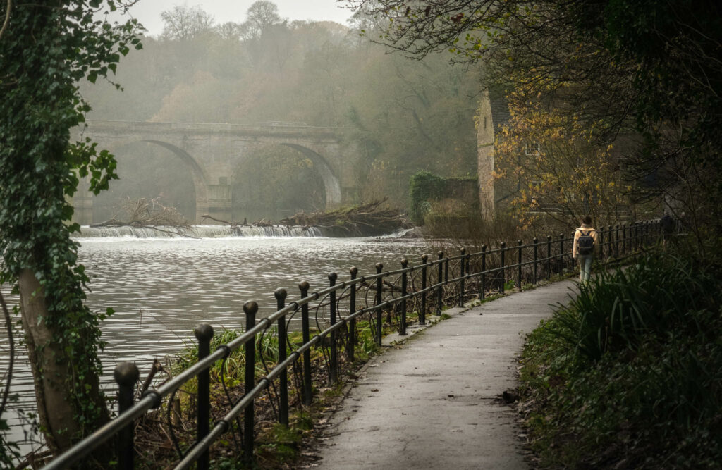 Woman walks pathway beside River Wear in Durham with Prebends Bridge in background