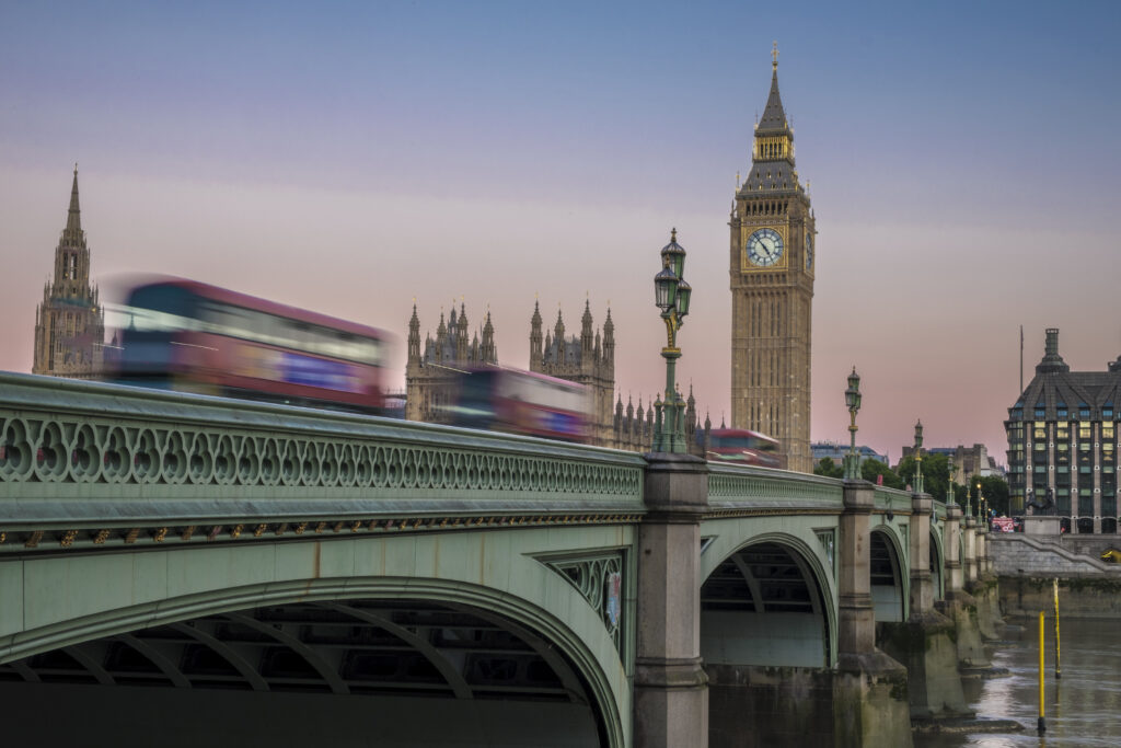 Three buses speed across Westminster Bridge with the houses of parliament behind