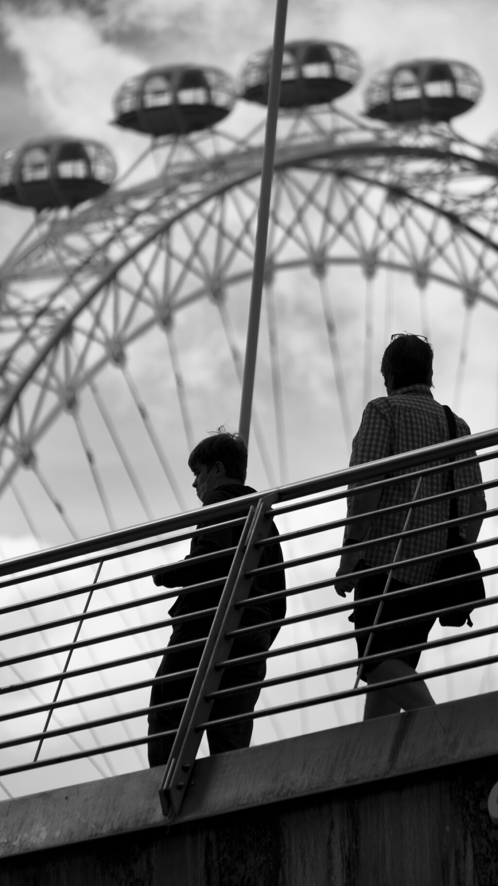 Two people walk across Hungerford Bridge with the London Eye behind. Shot in black and white 
