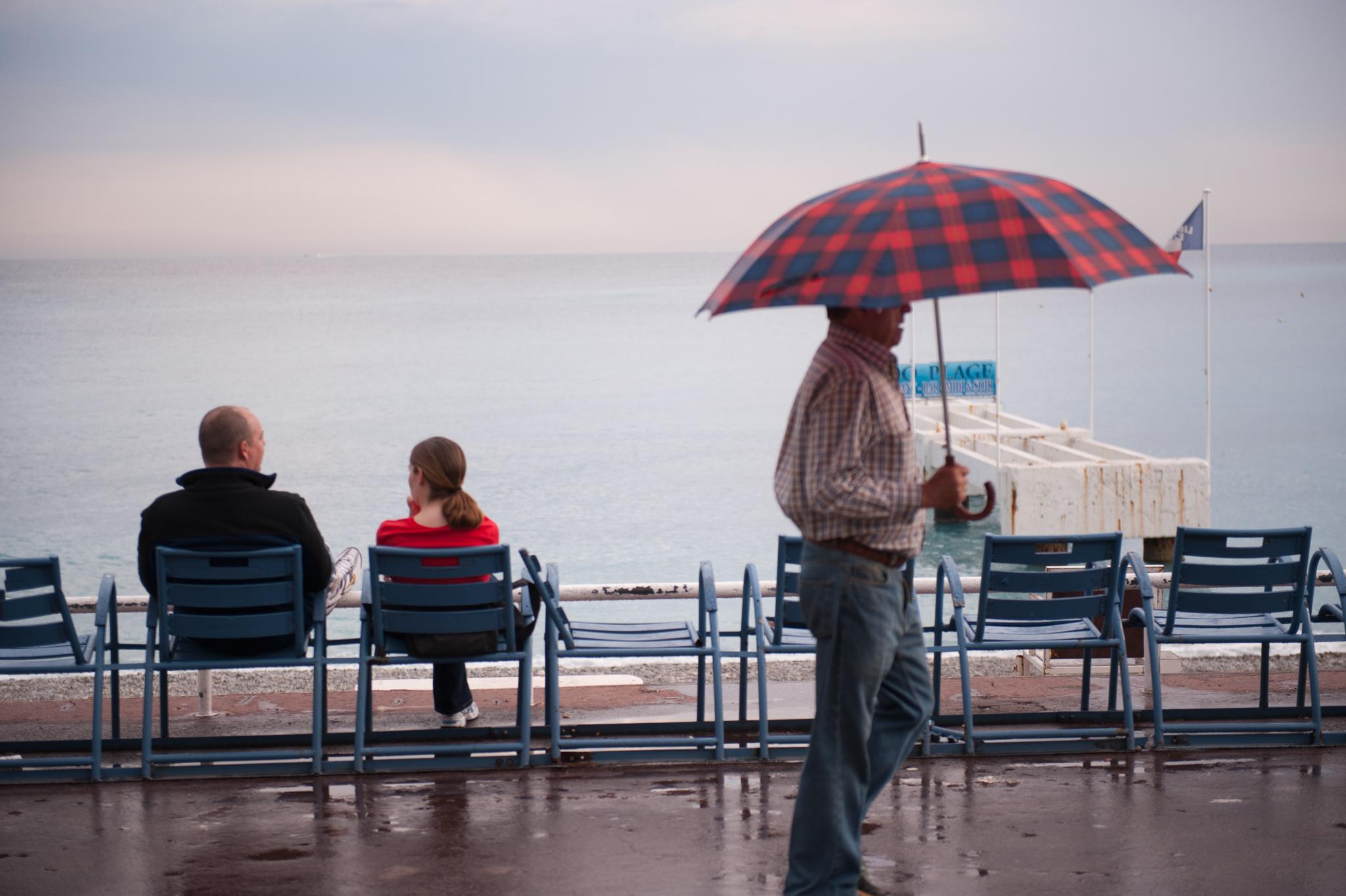Man with umbrella walks past two tourist on Promenade Des Anglaise in Nice France