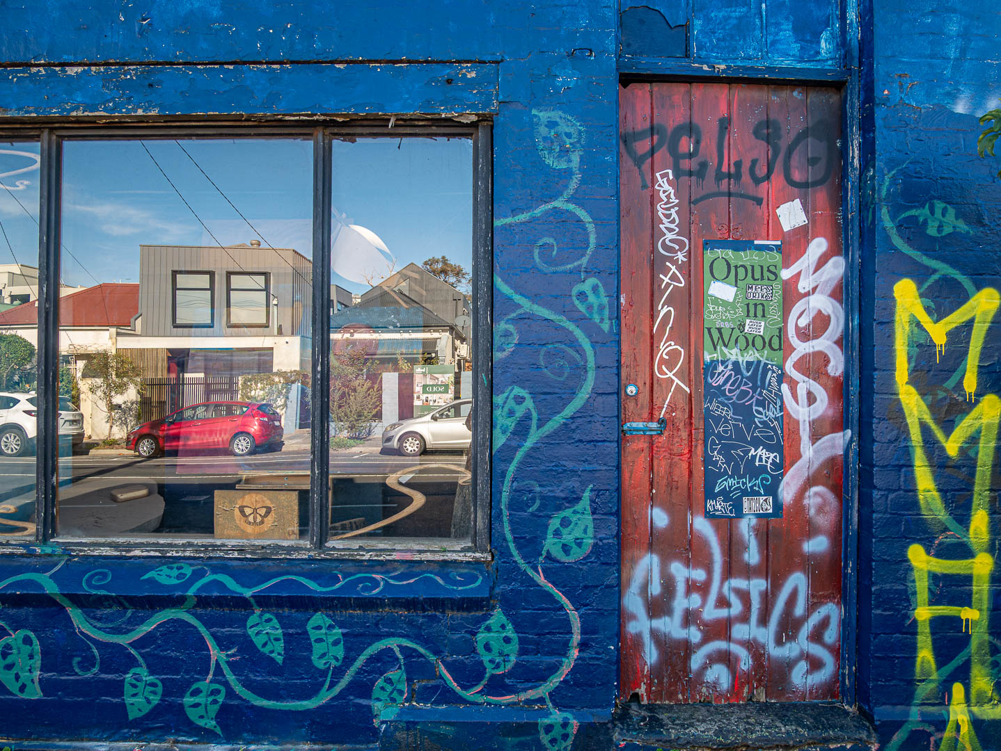 A colourful urban wall with a blue façade, red door, green paint and yellow graffiti, photographed by Anne Milton